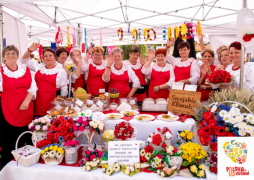 Grupa kobiet w białych bluzkach i czerwonych fartuchach. Na stole widoczne jedzenie i kwiaty
