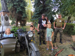 Grupa osób: dorośli i dzieci. Widoczny wojskowy motocykl 
