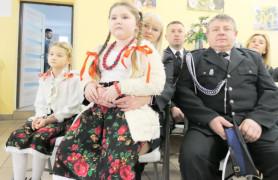 Dzieci w ludowych strojach i dorośli w mundurach strażackich siedzą obok siebie