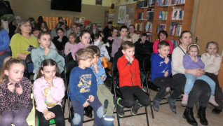 Dzieci i dorośli znajdują się w bibliotece. Wszyscy siedzą na krzesłach 