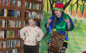 Kobieta w kolorowym ubraniu. Obok niej stoi dziewczynka w swetrze w serduszka. Teatralne przedstawienie  