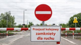 Widoczny biało-czerwony szlaban oraz znak zakaz wjazdu przy przejeździe kolejowym. Widoczna tablica informacyjna z napisem: Przejazd kolejowy zamknięty dla ruchu 
