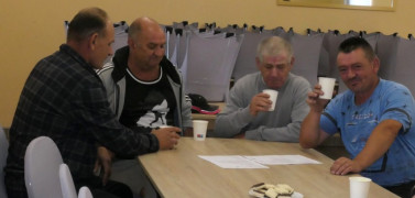 Czterej panowie siedzą przy stole. Panowie w dłoniach trzymają białe kubeczki. Uczestnicy warsztatów kulinarnych