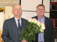 Dwaj panowie stoją obok siebie. Jeden z nich trzyma w ręku bukiet róż 