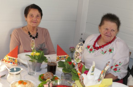 Dwie panie siedzą obok siebie przy stole, na którym znajduje się jedzenie i kwiaty
