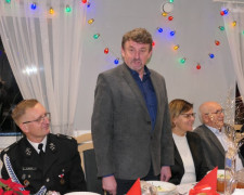 Spotkanie opłatkowe: osoby siedzą przy stole. Jedna z nich, mężczyzna stoi i przemawia 