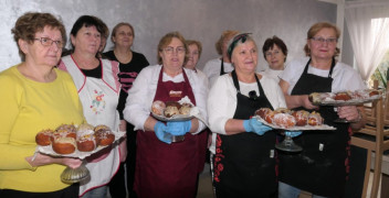 Grupa kobiet stoi obok siebie. Kobiety trzymają w dłoniach tace, na których znajdują się pączki