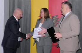 Dwaj mężczyźni i kobieta. Mężczyzna podaje rękę kobiecie. Osoby trzymają w dłoniach dokumenty
