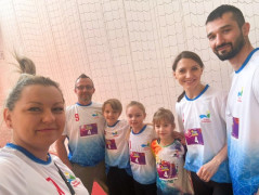 Dorośli i dzieci stoją obok siebie. Wszyscy są uśmiechnięci i ubrani w kolorowe koszulki