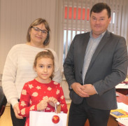 Kobieta, dziewczynka i mężczyzna stoją obok siebie. Dziewczynka trzyma w ręku upominkową torbę