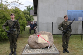 Obelisk przepasany biało-czerwoną wstążką. Obok obelisku stoją dwaj żołnierze 