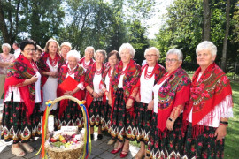 Grupa kobiet w kolorowych strojach. Przed kobietami stoi wieniec dożynkowy 