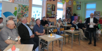 Grupa osób siedzi przy stolikach. Jeden z mężczyzn stoi i przemawia 