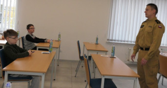 Dwaj chłopcy siedzą przy stolikach. Przed nimi stoi strażak