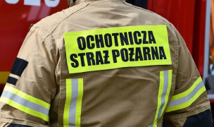 Mężczyzna stoi tyłem. Ubrany jest w strój strażaka. Na plecach widnieje napis: Ochotnicza Straż Pożarna 