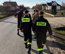 Strażacy idą drogą. Przed nimi widać domostwa