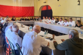 Grupa osób siedząca za stołem. Osoby te obradują. 