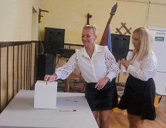 Dwie kobiety w białych bluzkach. Jedna z nich wrzuca do urny kartę do głosowania