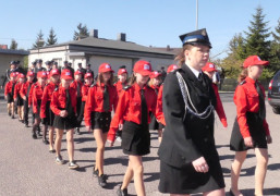 Defilada: widoczna kobieta w mundurze. Za kobietą idą dzieci w czerwonych koszulach 