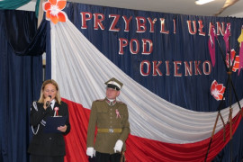 Kobieta i mężczyzna oboje w mundurach. Za nimi znajduje się biało - czerwona flaga