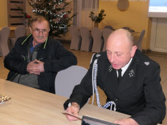 Dwóch mężczyzn siedzi za stołem. Jeden z nich ubrany jest w strażacki mundur 