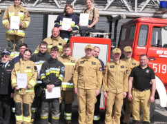 Grupa osób w strojach straży pożarnej stoi na tle wozu bojowego