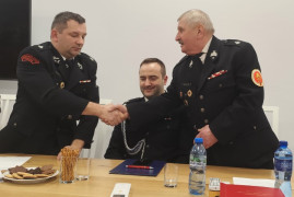 Trzej panowie w mundurach strażackich. Dwaj z nich podają sobie dłonie