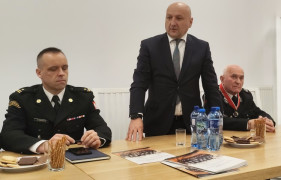 Trzech mężczyzn. Dwóch w mundurach strażaków, jeden z garniturze. Mężczyzna w garniturze stoi i przemawia