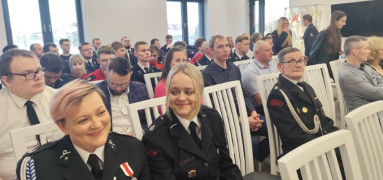 Grupa osób siedząca na krzesłach. Na pierwszym panie dwie uśmiechnięte kobiety w mundurach strażackich 