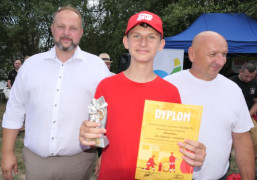 Dwaj mężczyźni w białych koszulach i chłopiec w czerwonej bluzce i czapce. Chłopiec trzyma statuetkę i dyplom 