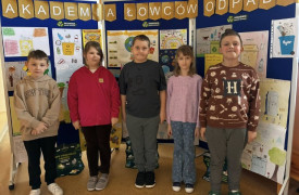 Uczniowie stoją przed ścianką, na której znajdują się prace plastyczne 