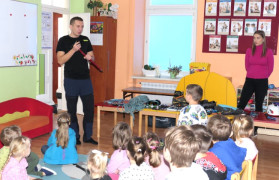 Grupa dzieci i mężczyzna. Mężczyzna prezentuje dzieciom sprzęt przeznaczony do wspinaczki górskiej 