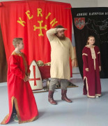 Mężczyzna i dwoje dzieci: dziewczynka i chłopiec stoją obok siebie. Wszyscy ubrani są w historyczne stroje. Trwa pokazowa lekcja dotycząca czasów pierwszych Piastów 