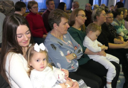 Osoby siedzą obok siebie. Na kolanach kobiety siedzi mała dziewczynka 