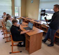 Uczniowie siedzą w szkolnych ławkach. Nagrywa ich mężczyzna 