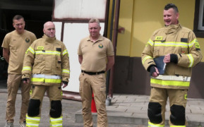 Czterej strażacy stoją obok siebie. Jeden z nich trzyma w dłoniach dokumenty 