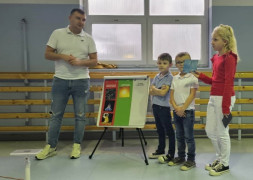 Trzy dziewczynki i mężczyzna stoją przy kolorowej tablicy. Mężczyzna tłumaczy dziewczynkom zasady powstawania prądu 