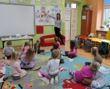 Dzieci siedzą na dywanie. Nauczycielka stoi przy tablicy. Dzieci słuchają tego co mówi im Pani