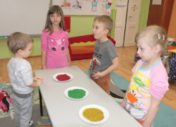 Przedszkolaki stoją przy stoliku, na którym znajdują się talerze z kolorowym brokatem 