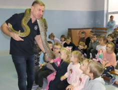 Dzieci siedzą obok siebie na ławce. Mężczyzna trzyma węża, opowiada o nim i pozwala dzieciakom dotknąć go
