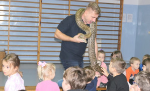 Sala gimnastyczna w szkole. Dzieci siedzą na długiej ławce. Mężczyzna trzyma węża na szyi, opowiada im o gadach. Dzieci mają możliwość dotknięcia węży