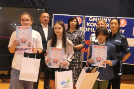 Grupa osób stoi obok siebie. Dziewczynki i chłopiec trzymają w dłoniach dyplomy 