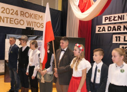 Grupa dzieci - uczestnicy szkolnej akademii. Jeden z chłopców trzema biało-czerwoną flagę 