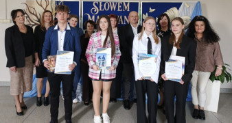 Grupa osób dorośli i nastolatkowie stoją obok siebie. Nastolatkowe trzymają w dłoniach dyplomy 