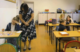 Uczniowie siedzą w ławkach i rozwiązują zadania. Obok jednego ucznia stoi kobieta 