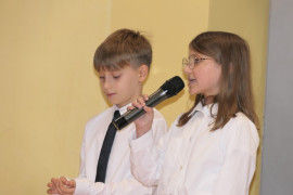 Dziewczynka i chłopiec podczas szkolnej akademii. Dziewczynka trzyma w ręku mikrofon 