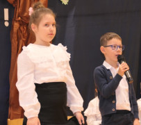 Dziewczynka i chłopiec w galowych strojach. Chłopiec trzyma w ręku mikrofon 