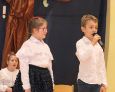 Dziewczynka i chłopiec w galowych strojach. Chłopiec trzyma w ręku mikrofon 