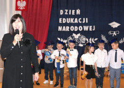 Grupa uczniów w galowych strojach. Przed uczniami stoi kobieta, która w ręku trzyma mikrofon 