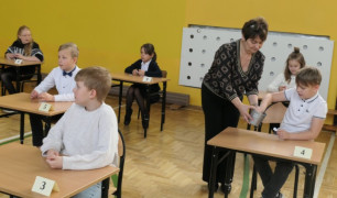 Dzieci siedzą w ławkach. Przy jednym z chłopców stoi kobieta 